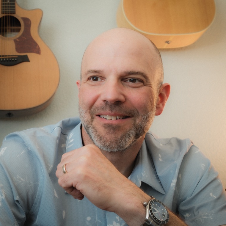 Scott Elias, smiling, with two acoustic guitars on the wall behind him.